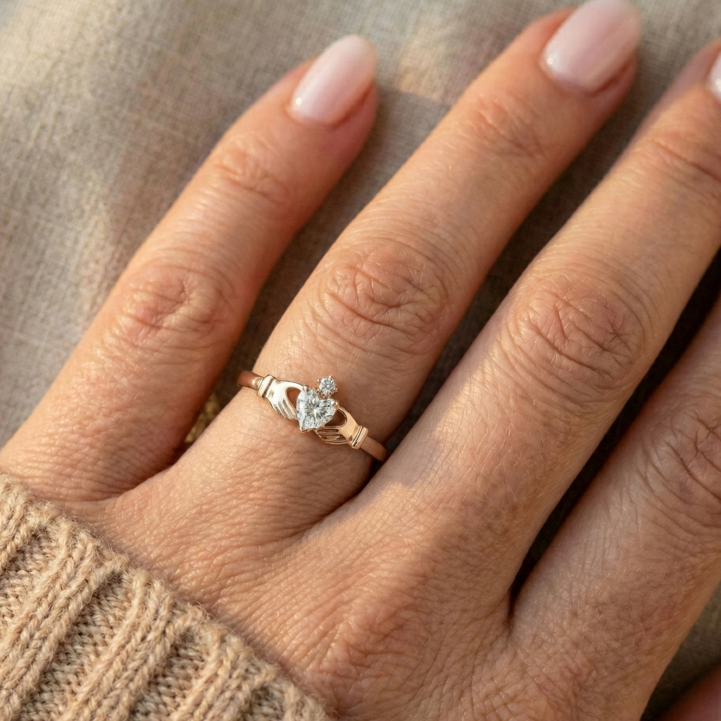 Hand wearing a rose gold claddagh ring with a diamond on a neutral background